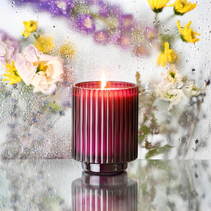 Editorial Shot of the lit Amélie - Lavender 12.3oz candle placed in the center of a rainy mirror scene, with several bunches of flowers arranged behind the candle