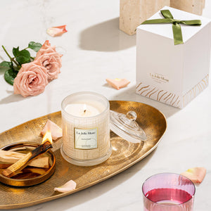 Photo shot of a lit Roesia - Santal Rosé 10oz candle placed on a dinner plate, surrounded by lit wooden sticks, roses, and candle packaging