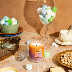 Photo shot of a litRoesia - Zest Pistachio Macaron 10oz candle placed in the middle of the scene, surrounded by macarons, pistachios, cheese, and a set of cutlery.