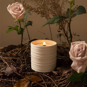  Photo shot of a lit Lucienne - Wild Rose 7.1 oz candle surrounded by branches, dried leaves, and three roses inserted