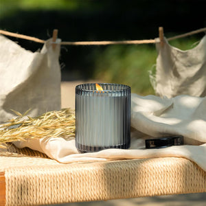 Editorial shot of outdoor scene: Sunlight streaming down, illuminating the lit Amélie - Breezy Linen 12.3oz candle, placed on a stool with a cotton-linen cloth underneath. Beside the candle, several bundles of wheat are arranged, while behind the stool, two pieces of cotton cloth are hanging to dry.