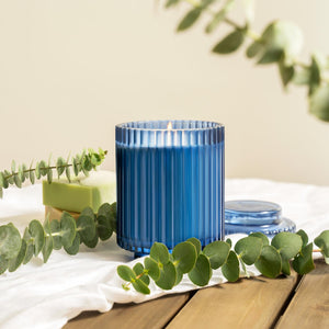 Editorial shot of the lit Amélie - Eucalyptus & Rosemary 12.3oz candle placed in the center of a wooden table, with a white tablecloth underneath, surrounded by several potted plants