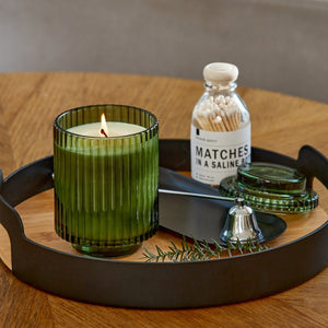Editorial Shot of Lit Amélie Fir & Cedarwood 12.3oz candle placed on a plate, which sits atop a wooden table. Positioned to the right of the candle are a bottle, a bell, and a sprig of fir.