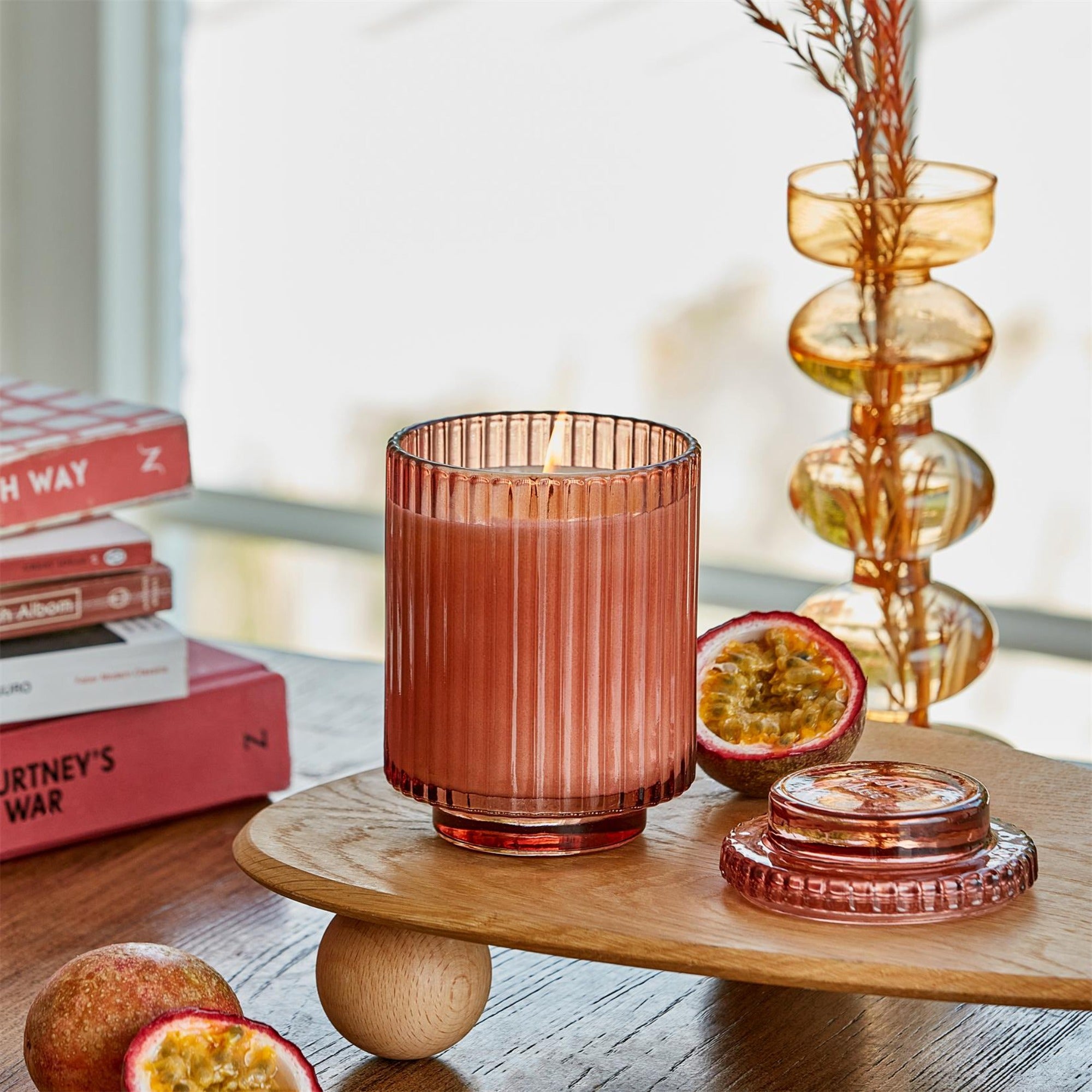 Editorial Shot of lit Amélie - Passionfruit Peony 12.3oz candle  placed to the left of a small table, surrounded by passion fruits, a stack of books, and a vase