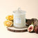 Photo shot of a lit Roesia - Wild Rose 9.9oz candle placed on a wooden board, surrounded by yellow roses and figs - view 2