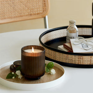 Editorial Shot of Lit Amélie Wood Jasmine 12.3oz candle placed on the left side of the table with three jasmine flowers surrounding it. Next to it is a bamboo woven plate, containing eyeglasses, a bottle of cotton swabs, and several books.