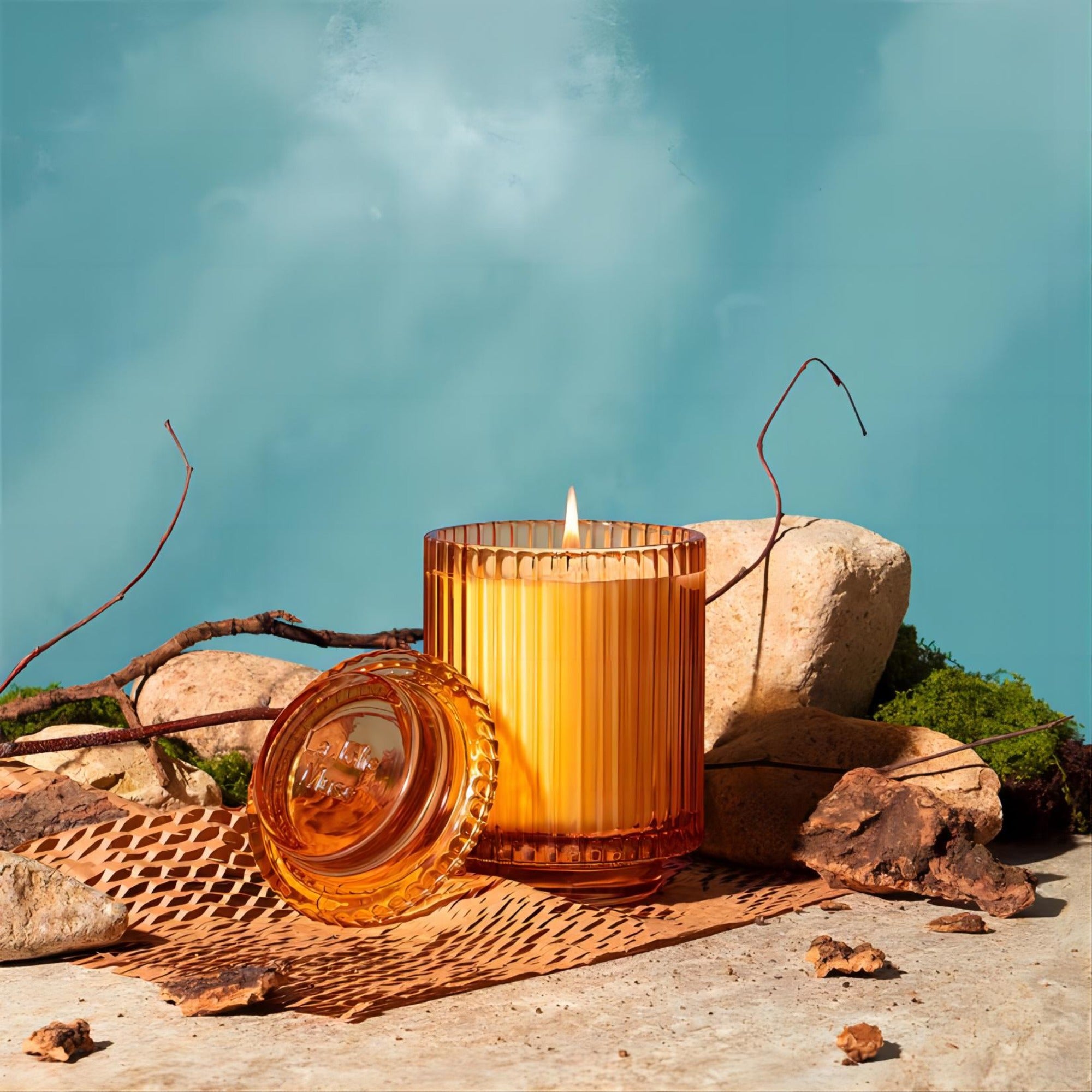 Editorial Shot of a lit Amélie - Sandalwood & Patchouli candle at the center, surrounded by stones, grass, and branches.