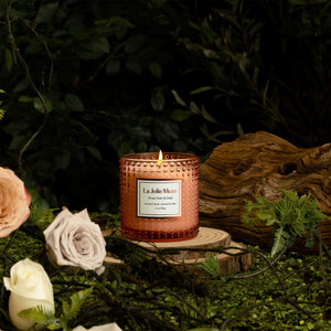 Photo shot of a lit Marvella - Rose Noir & Oud 10oz candle placed on a tree stump, surrounded by plants, roses, and branches
