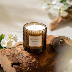Photo shot of a lit Marvella - Woody Jasmine 10oz candle placed on a wooden stump, with several jasmine plants nearby
