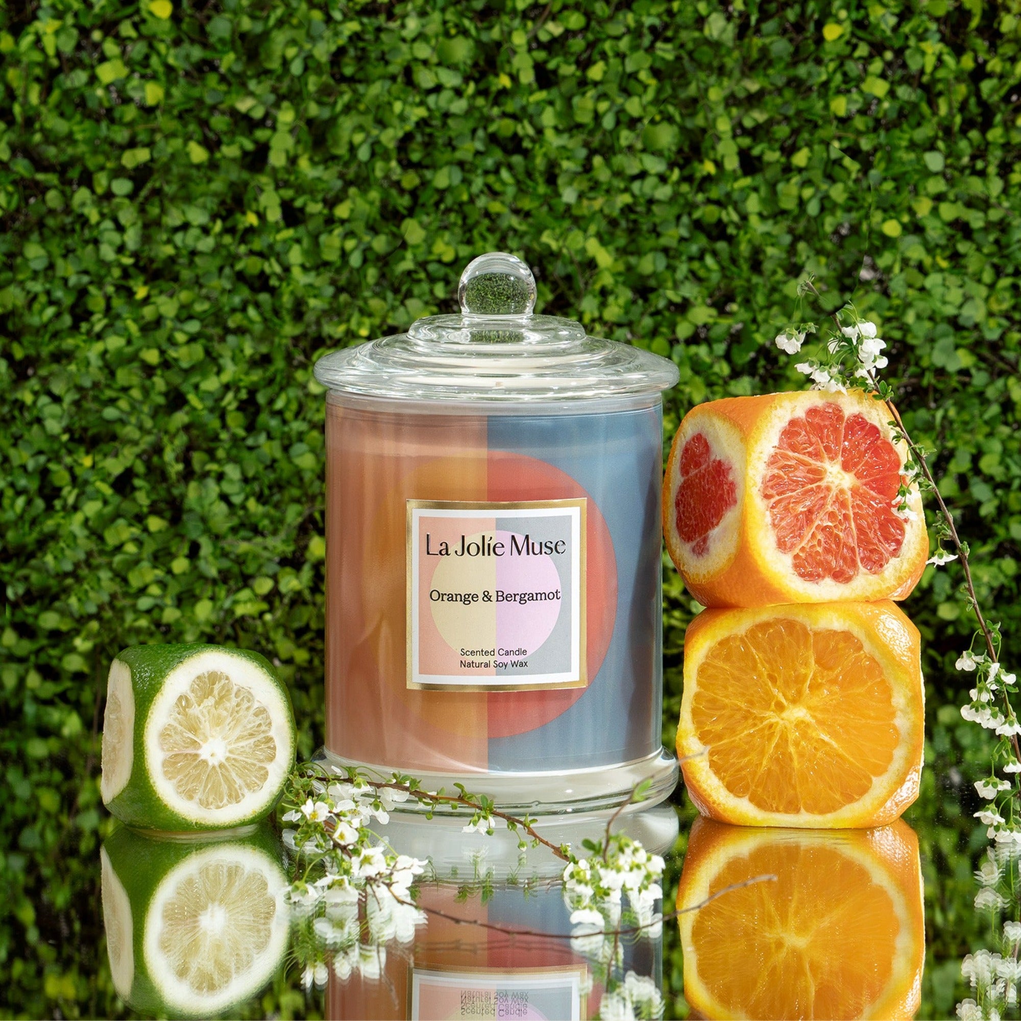 Photo shot of a Roesia - Orange & Bergamot 9.9oz candle placed in the middle, with lime on the left and orange and grapefruit on the right