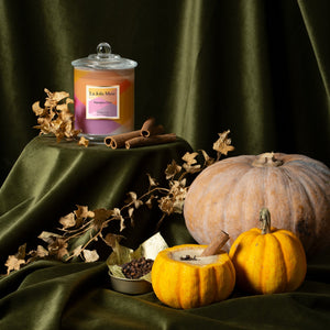 Photo shot of Roesia - Pumpkin Chai 9.9 oz candle placed on a pedestal, with cinnamon on the right and two pumpkins alongside a bowl of cloves