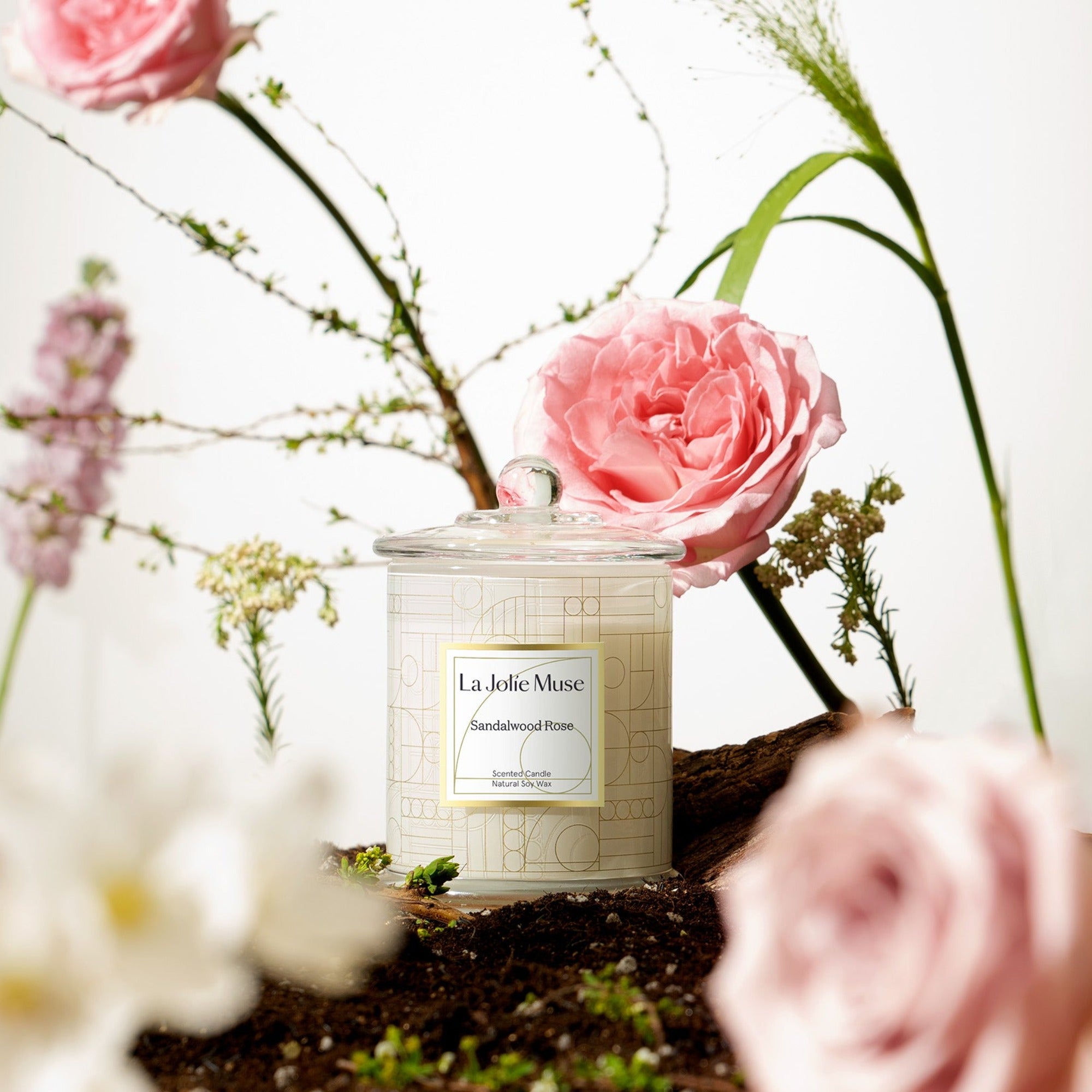 Photo shot of a lit Roesia - Sandalwood Rose 9.9oz candle placed in the middle of the ground, surrounded by planted roses