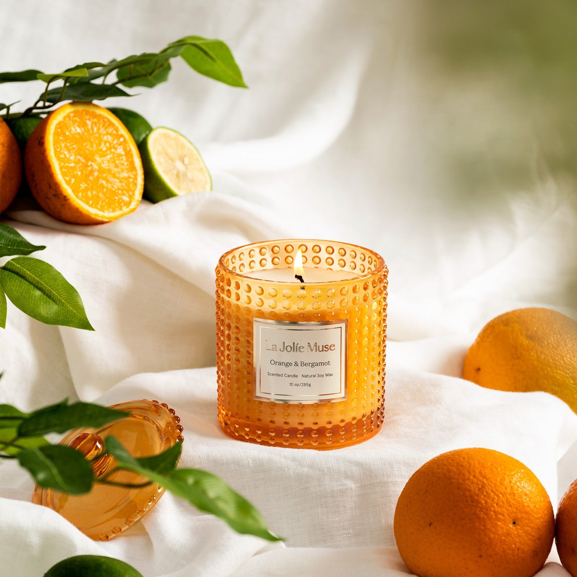 Photo shot of a lit Marvella - Orange & Bergamot 10oz candle placed on a tablecloth, surrounded by citrus fruits.