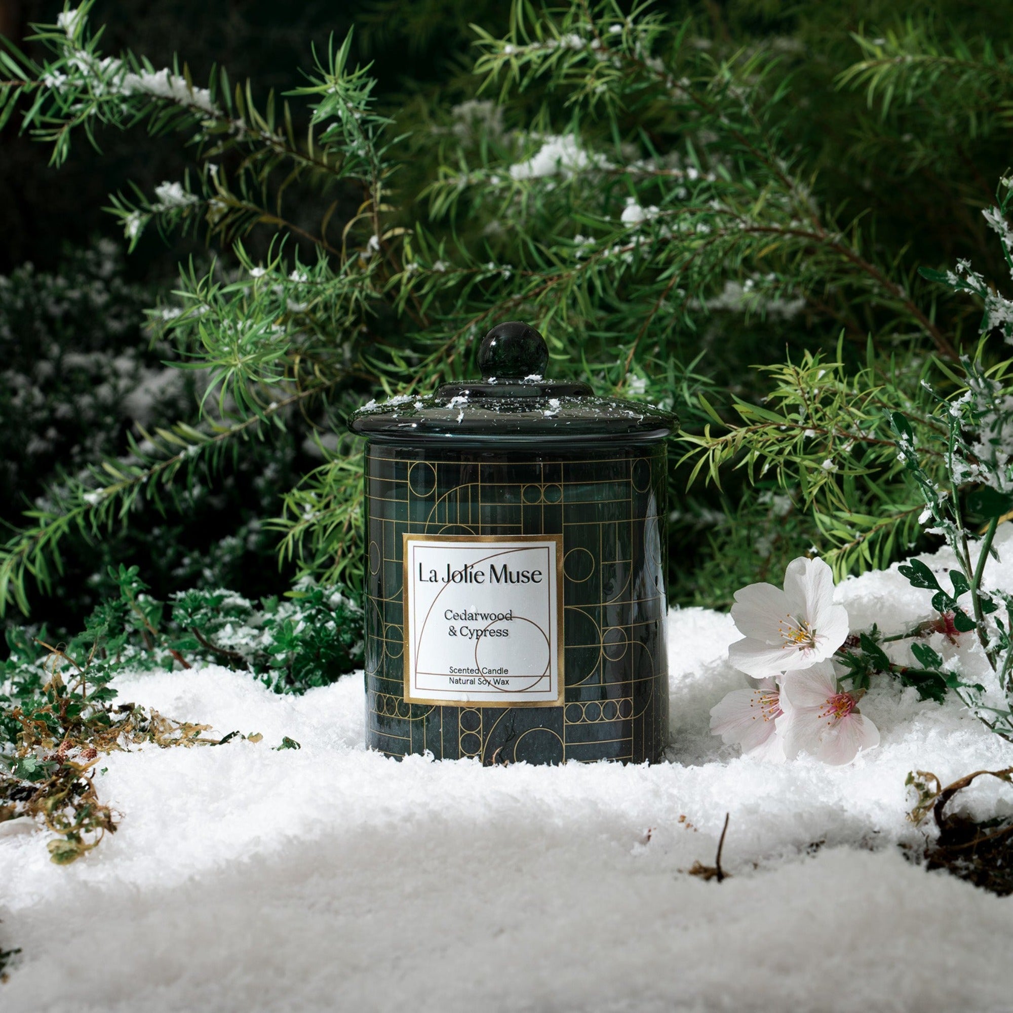 Photo shot of a Roesia - Pomegranate & Pine 10oz candle placed in artificial snow, surrounded by pine needles and flowers