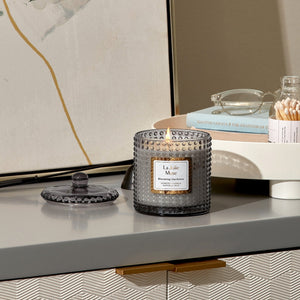 Photo shot of a lit Marvella - Blooming Gardenia 10oz candle placed on a cabinet, with glasses, books, and a jar of cotton swabs on the right