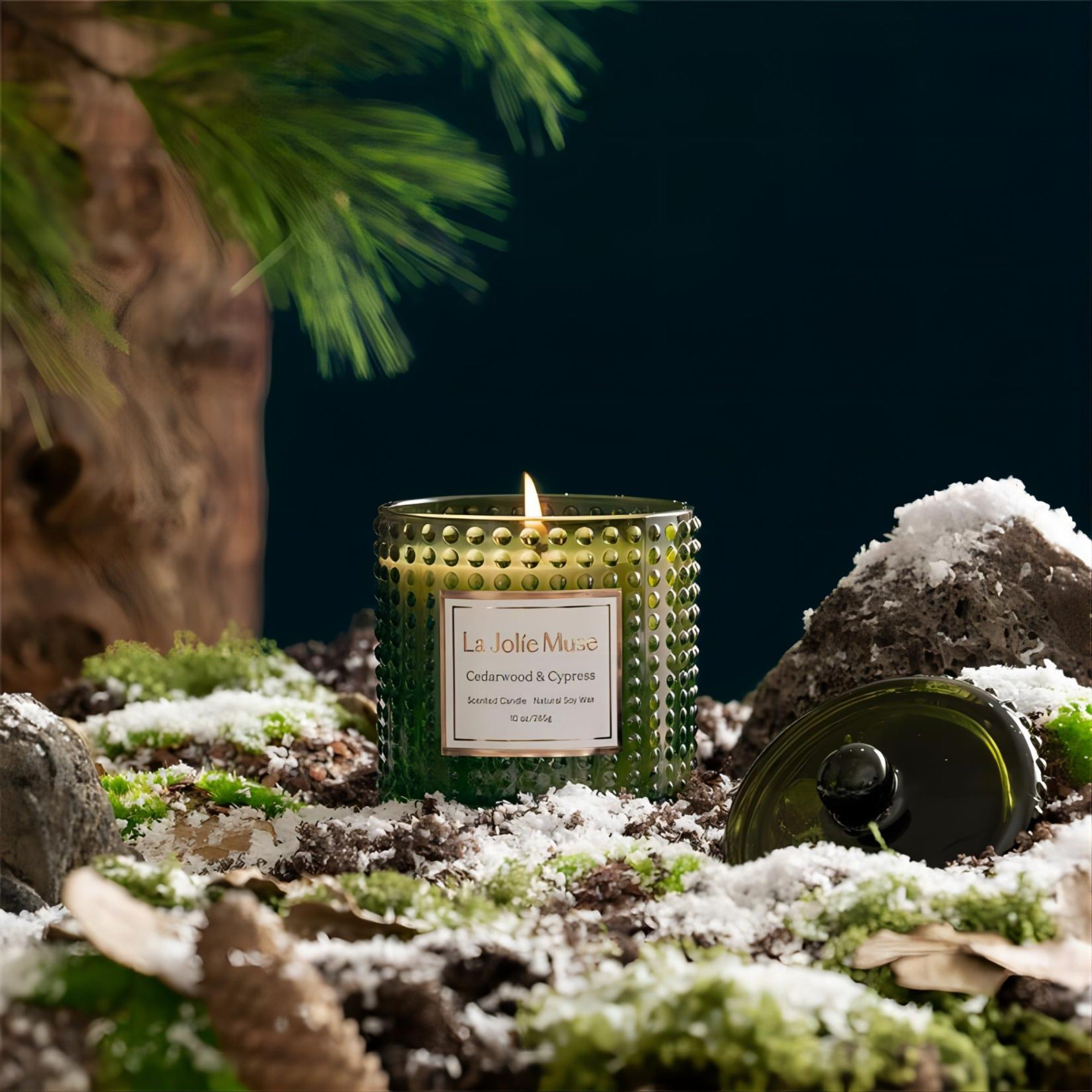  Photo shot of a lit Marvella - Cypress & Cedarwood 10oz candle placed on grass covered with artificial snow, surrounded by scattered stones, pine cones, and leaves