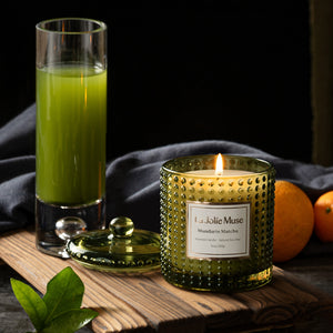  Photo shot of a lit Marvella - Mandarin Matcha 10oz candle placed on a wooden board, surrounded by a cup of matcha beverage, mandarins, and leaves