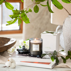 Photo shot of a lit Roesia - Linen Cotton Oasis 10oz candle placed on a table, with a tablecloth spread on the table, along with candle packaging, cotton, books, and plants.