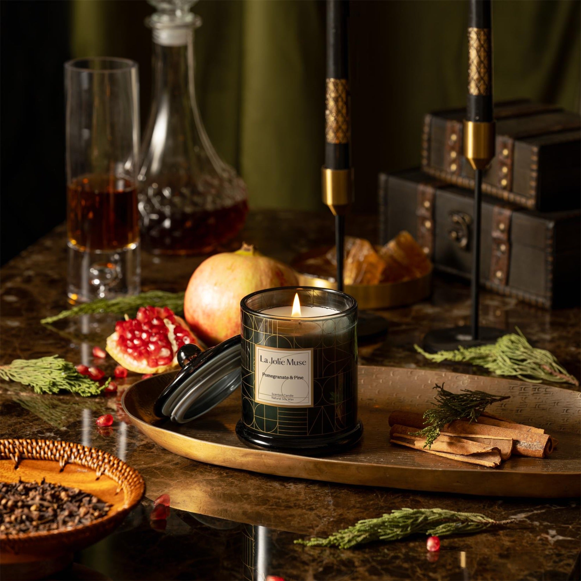 Photo shot of a lit Roesia - Pomegranate & Pine 10oz candle placed on a table, accompanied by pomegranates, pine, a leather storage box, wine, and cloves