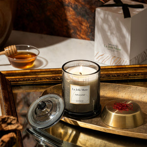 Photo shot of a lit Roesia Saffron & Oud 10oz candle placed on a metal plate, with a mirror underneath, surrounded by honey, saffron, and candle packaging.