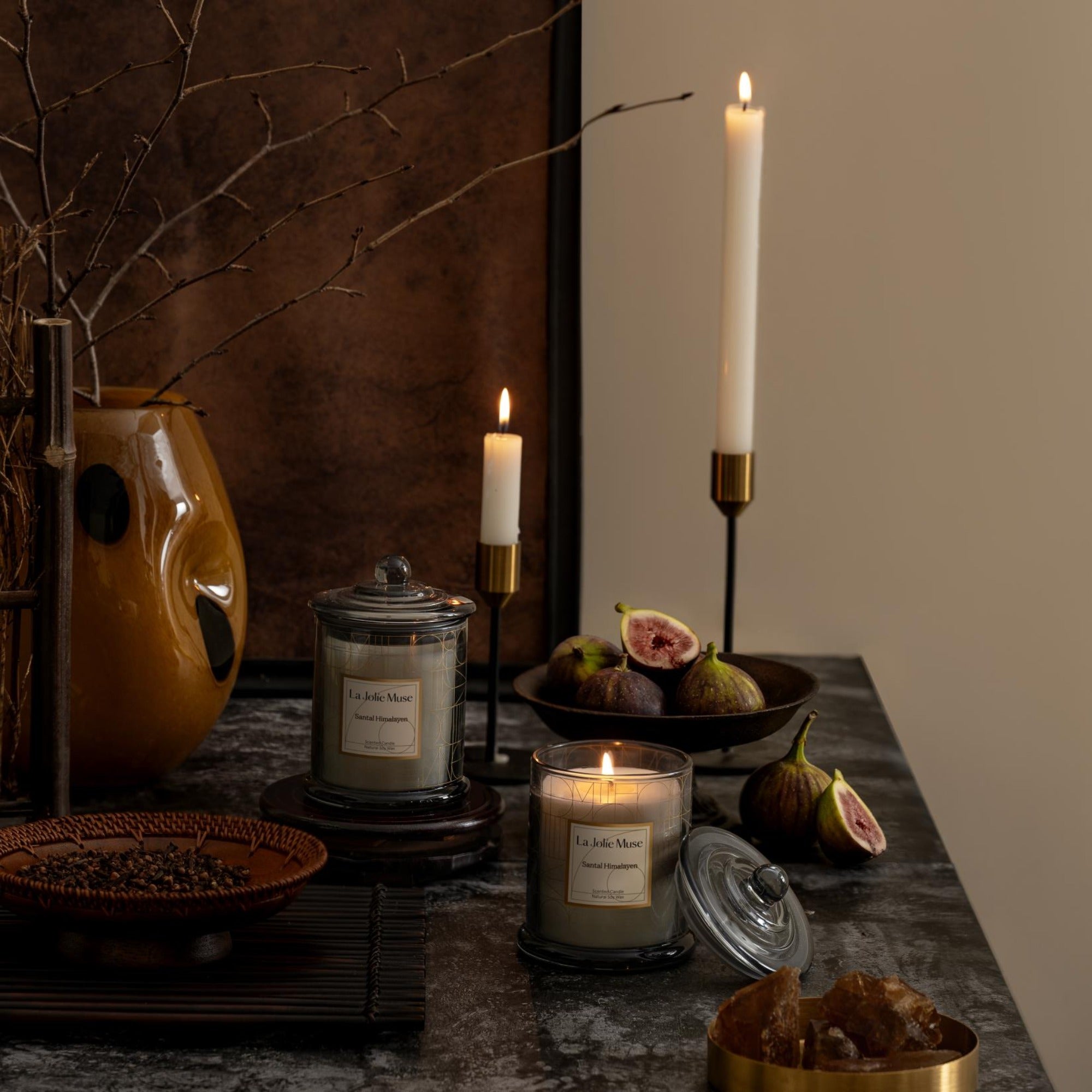  Photo shot of lit Roesia - Santal Himalayen 10oz candles placed on a marble table, surrounded by figs, candleholders, and a vase