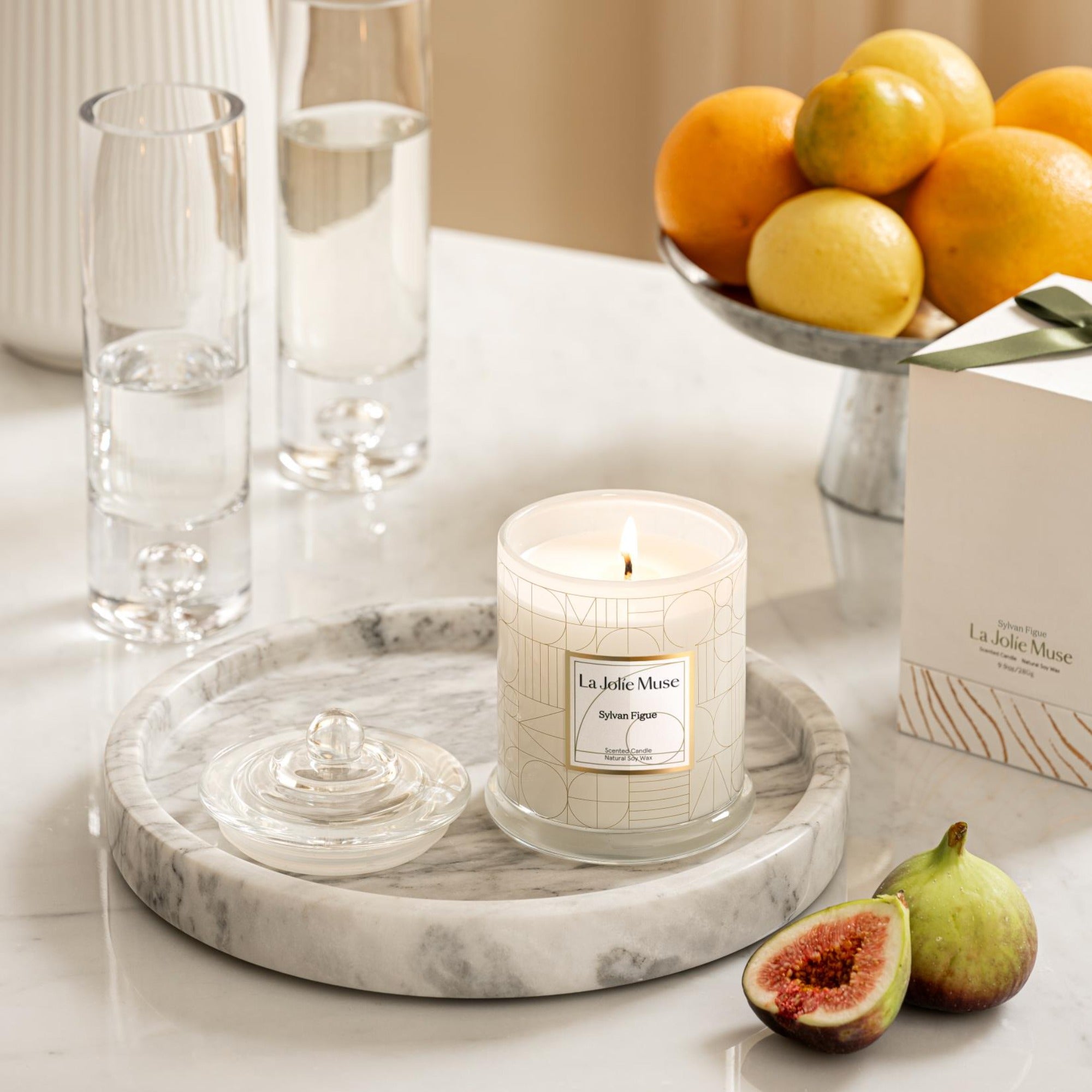 Photo shot of a lit Roesia - Sylvan Figue 10oz candle placed on a marble tray, surrounded by glasses, oranges, figs, and candle packaging