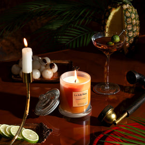 Photo shot of a lit Roesia - Zest Tahitian Lychee 10oz candle placed in the center, surrounded by a pipe, fruits, and a cocktail