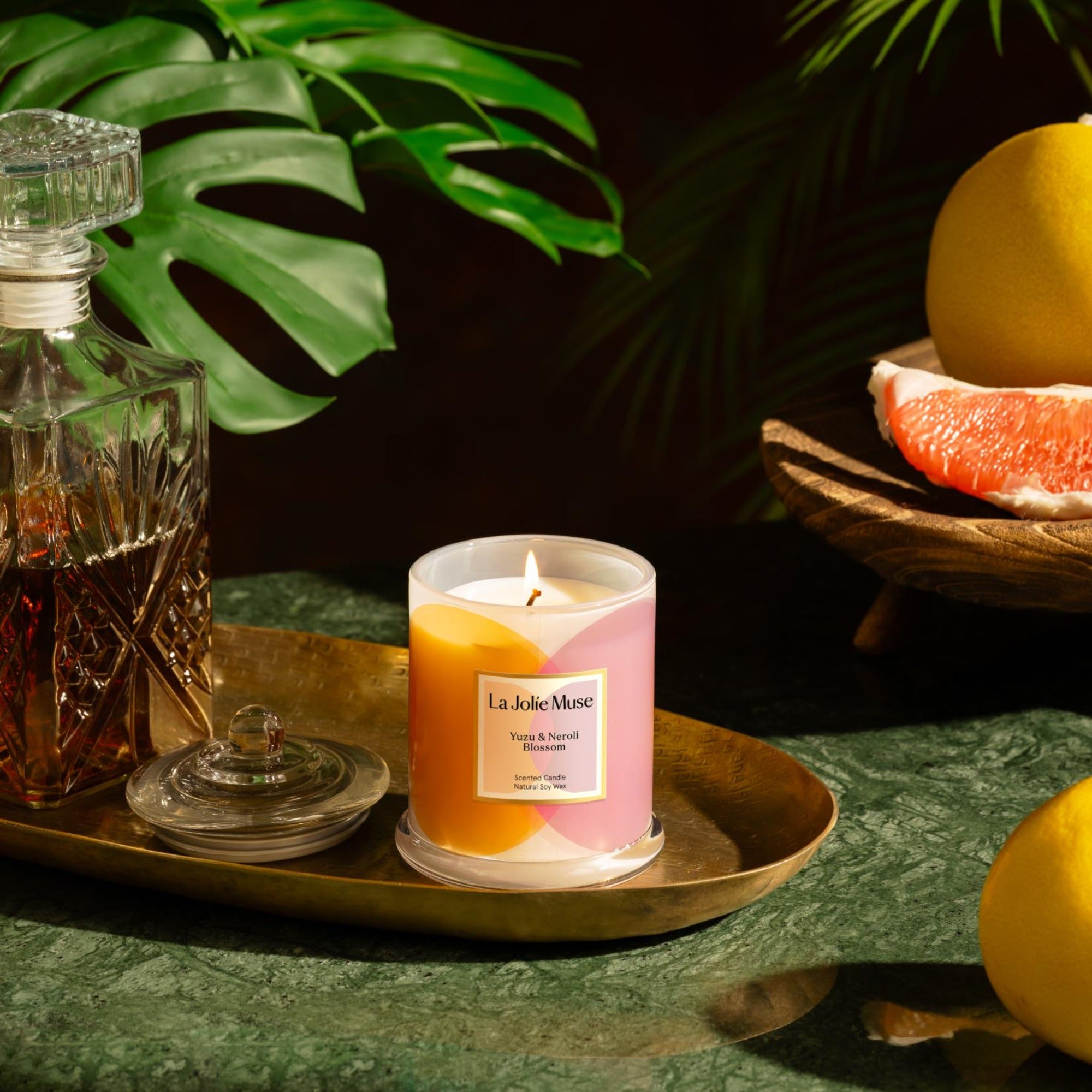 Photo shot of a lit Roesia - Zest Yuzu & Neroli Blossom 10oz candle placed on a wooden tray, with a bottle of wine on the left and yuzu on the right