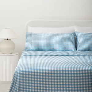 Paulina Blue Check Picot Edge 200 count cotton sheet set on a fully made bed. White and blue gingham duvet and two white pillows and two white and blue gingham pillows next to side table with lamp.