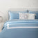 Paulina Sky Blue 200 thread count combed cotton duvet set on a fully made bed next to side table with plant. Gray and sky blue duvet, two sky blue and white pillows, two blue gingham pillows, and two fluffy white accent pillows.