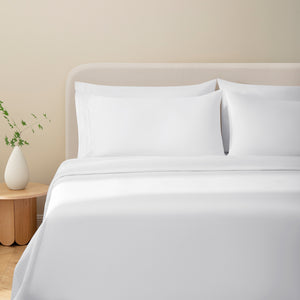 Sofia Classic White Double Satin Stitched Cuff 300 thread count cotton bed sheet set on a fully made bed. White cotton duvet and four white pillows next to side table with plant.