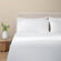 Camille Classic White sheet set on a fully made bed. White duvet and four white pillows  next to side table with plant.