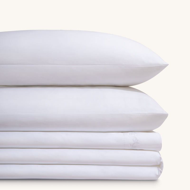 Nina Classic White 600 thread count cotton bed sheet set. Two white embroidered pillows stacked on folded white embroidered sheet set.