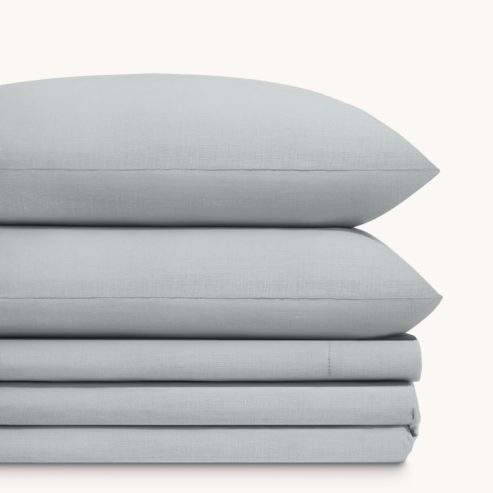Olivia Foggy gray linen bed sheet set. Two foggy gray linen pillows stacked on folded foggy gray linen sheet set.