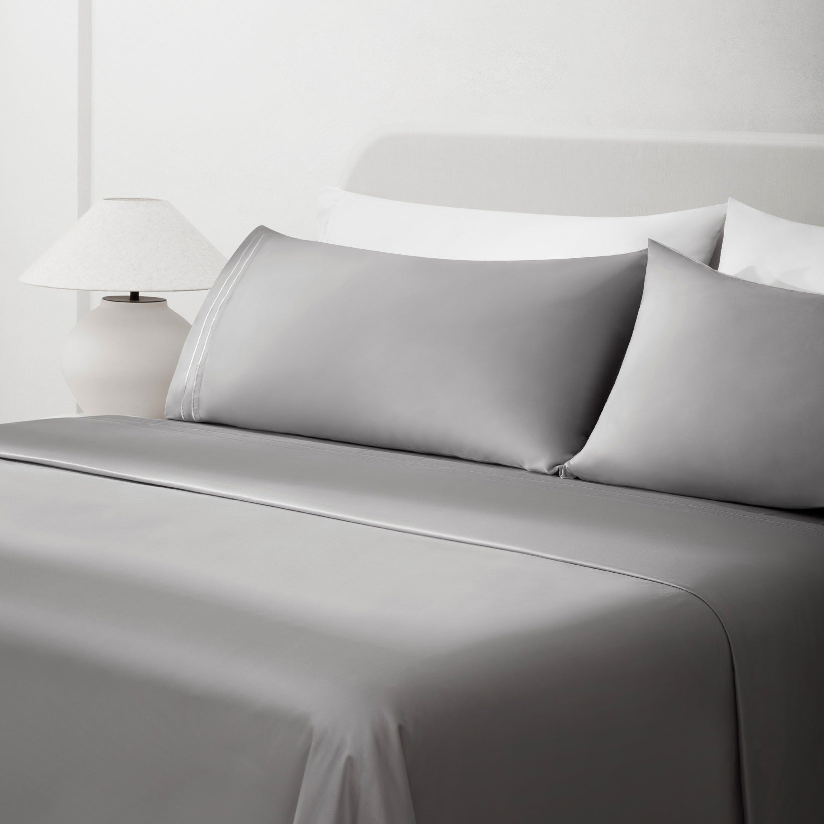 Sofia Foggy Gray Double Satin Stitched Cuff 300 thread count cotton bed sheet set. Gray pillows and gray cotton sheets on bed next to side table and lamp.
