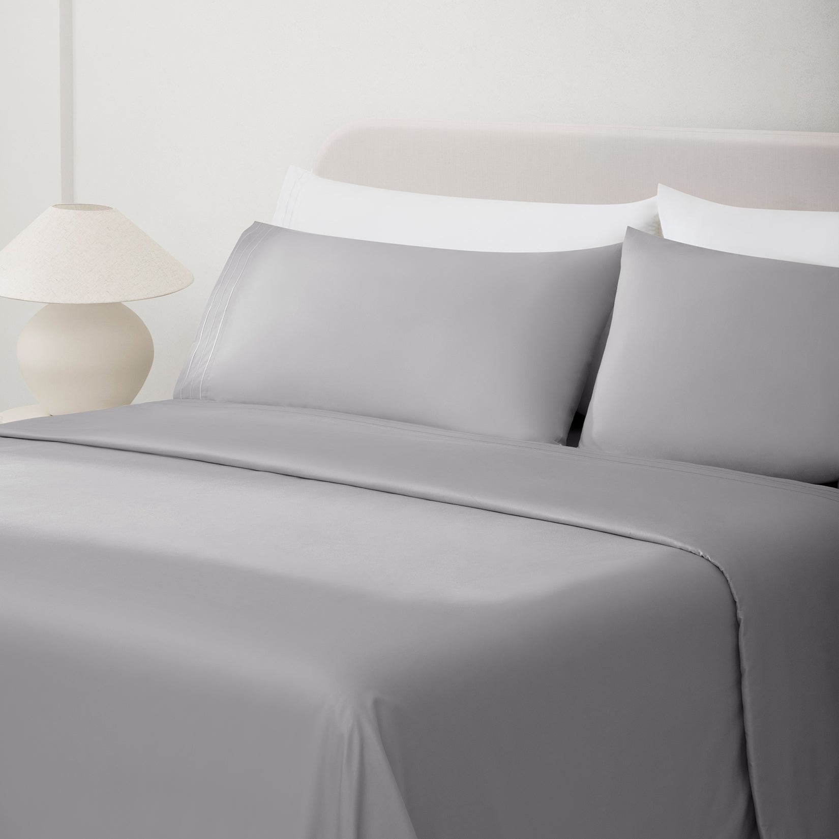Paulina Foggy Triple Satin Stitched Cuff 200 thread count cotton bed sheet set. Gray pillows and gray cotton sheets on bed next to side table and lamp.