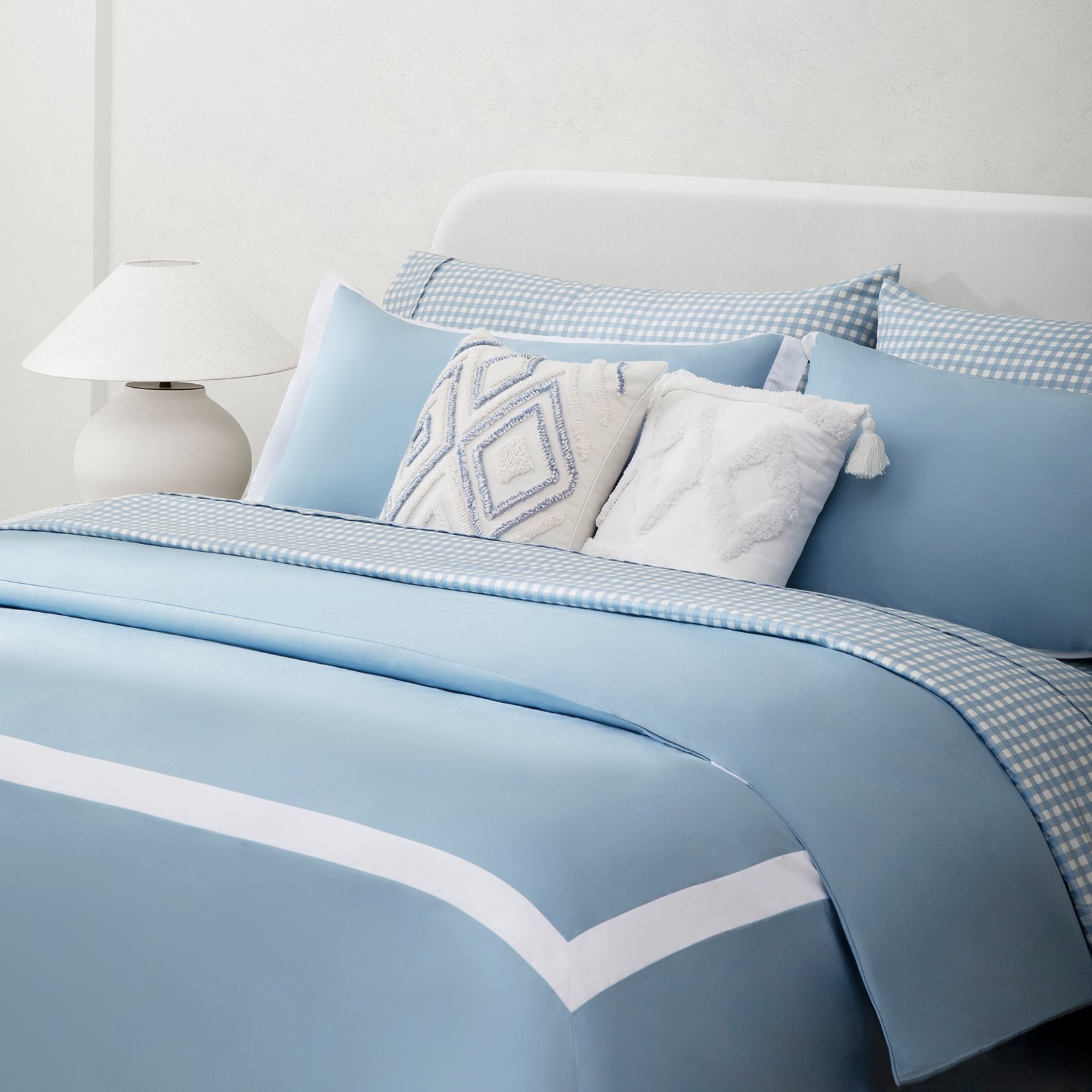 Paulina Sky Blue 200 thread count combed cotton duvet set. Sky blue and white pillows and duvet on sky blue gingham bed sheets with fluffy accent pillows next to side table and lamp.