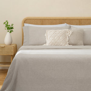 Olivia natural taupe linen sheet set on a fully made bed next to side table with plant and accent pillows. Natural taupe duvet with two white pillows, two natural taupe pillows.