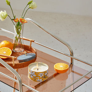 A burning candle is displayed on a transparent tea stand with fresh oranges and a vase of flowers.