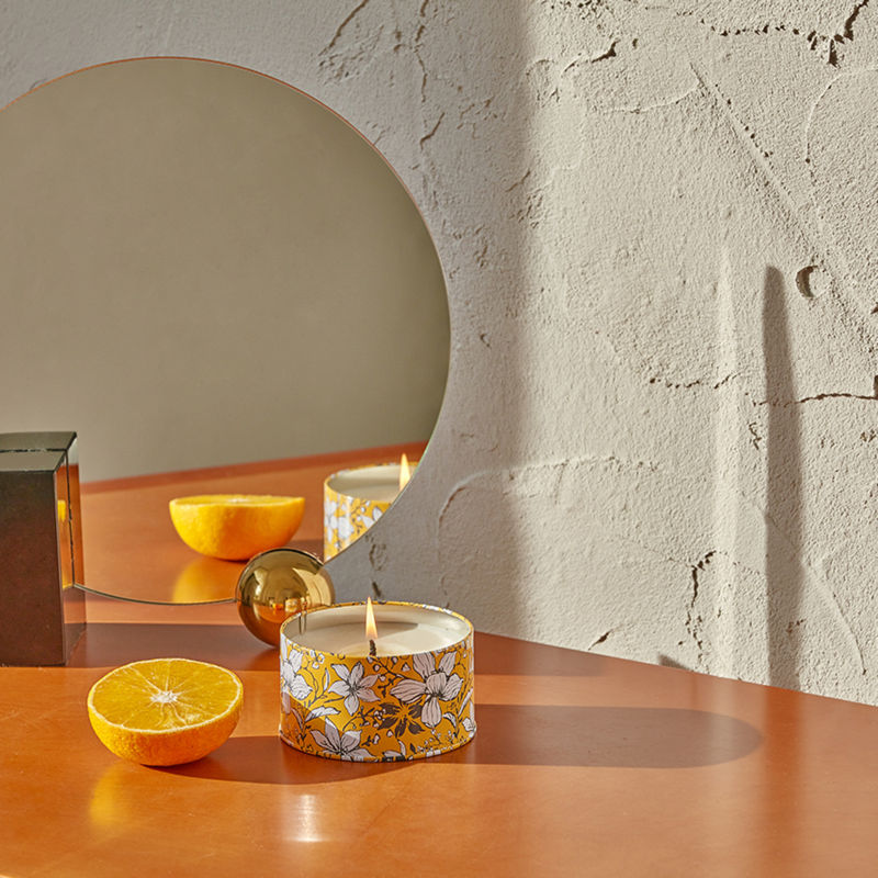 A jar of burning candle is displayed with half an orange, in front of a round mirror.