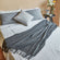 An Angie smoky gray faux fleece fringed throw blanket is displayed on a bed with three pillows.