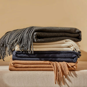 Four colors of folded Angie faux fleece fringed throw blankets are displayed on a wooden shelf in front of a window.
