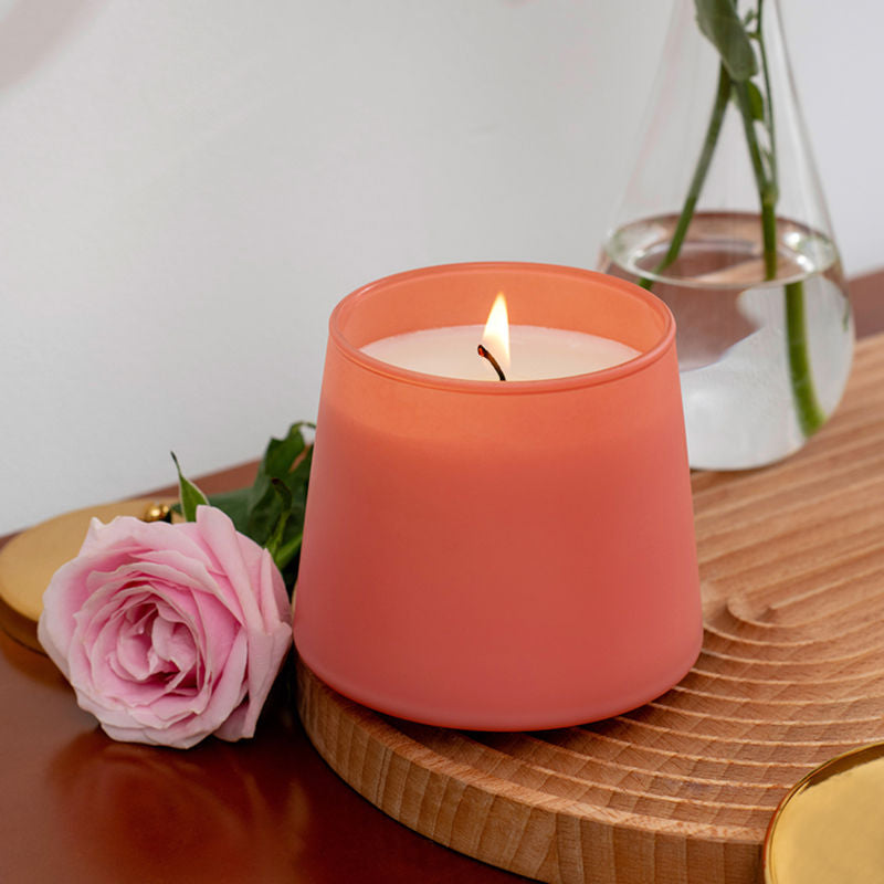 A jar of burning candle is placed on a wooden tray with a pink rose beside.