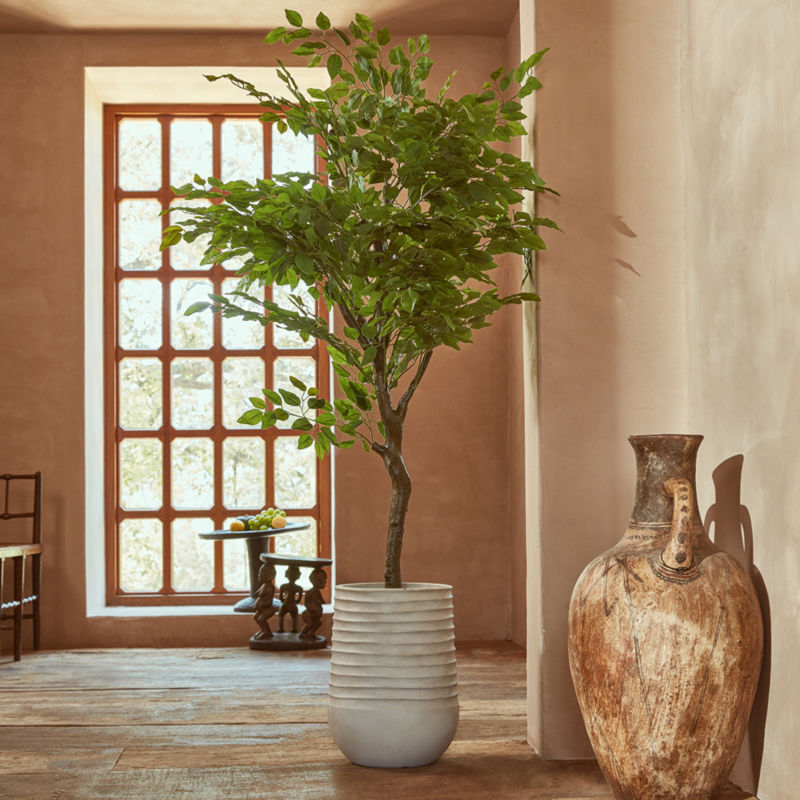 A picture staging the artificial ficus tree in a large white pot. Behind the tree, there are tables, a chair and a window.