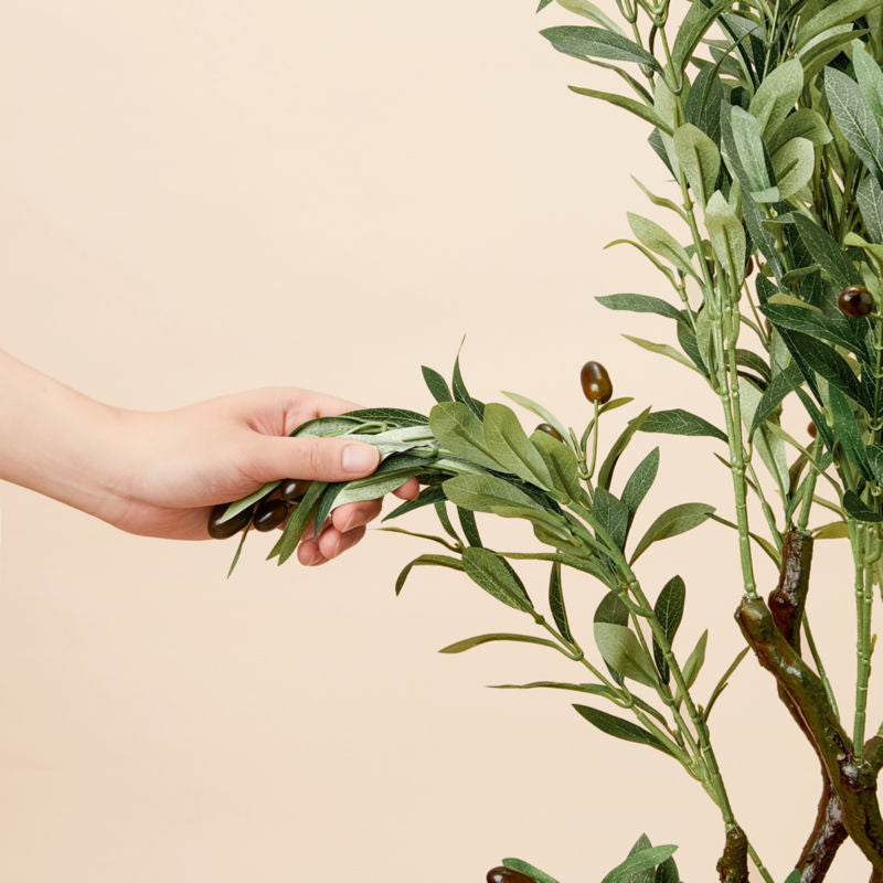 A hand grasps a branch and the leaves on an artificial olive tree, bending it to show it is easily styleable.