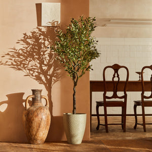 A faux olive tree is planted in a gray pottery and displayed near a brown pottery.