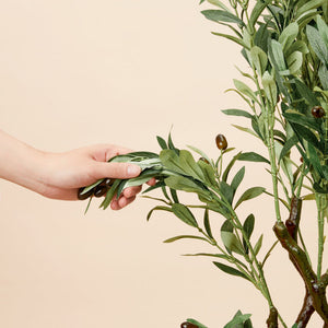 A hand grasps the branch of an artificial olive tree, showing its flexible and adjustable components.