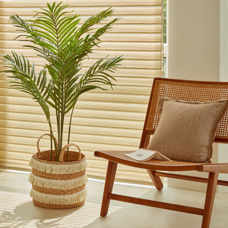 The artificial palm tree is placed in a basket near a curtained window and a chair.