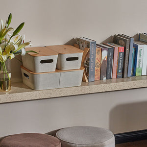 A set of three beige bamboo utility baskets baskets are set on a marble wall shelf with a row of books beside.