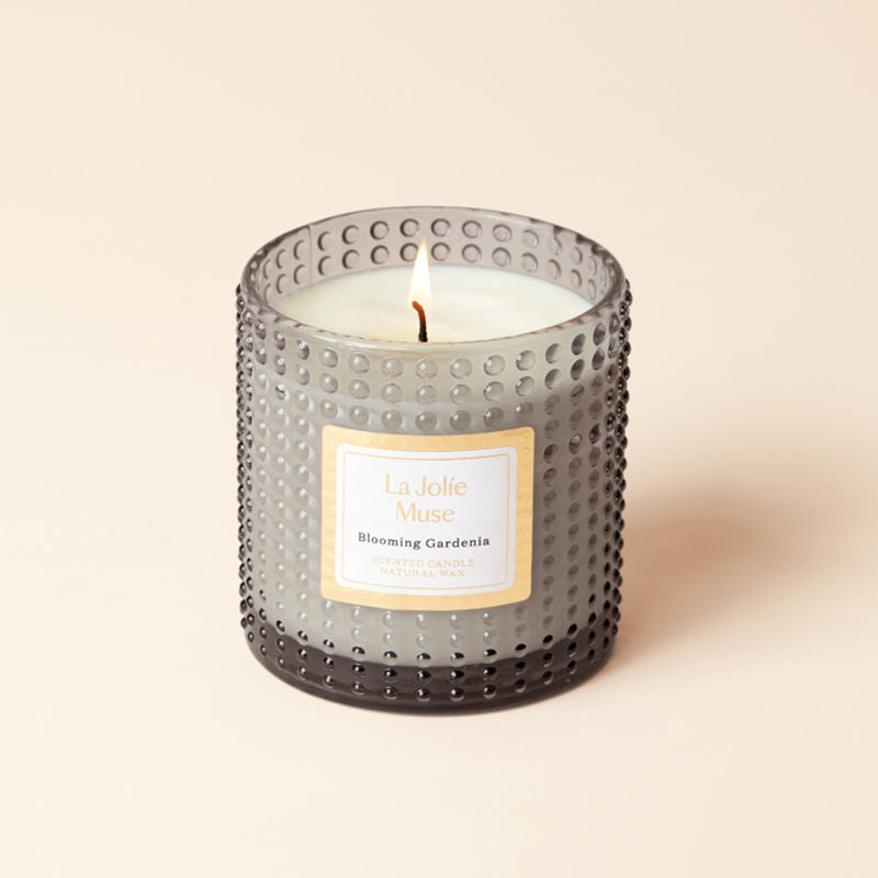 A full view of a burning  Blooming Gardenia candle, placed in a grey glass jar. 